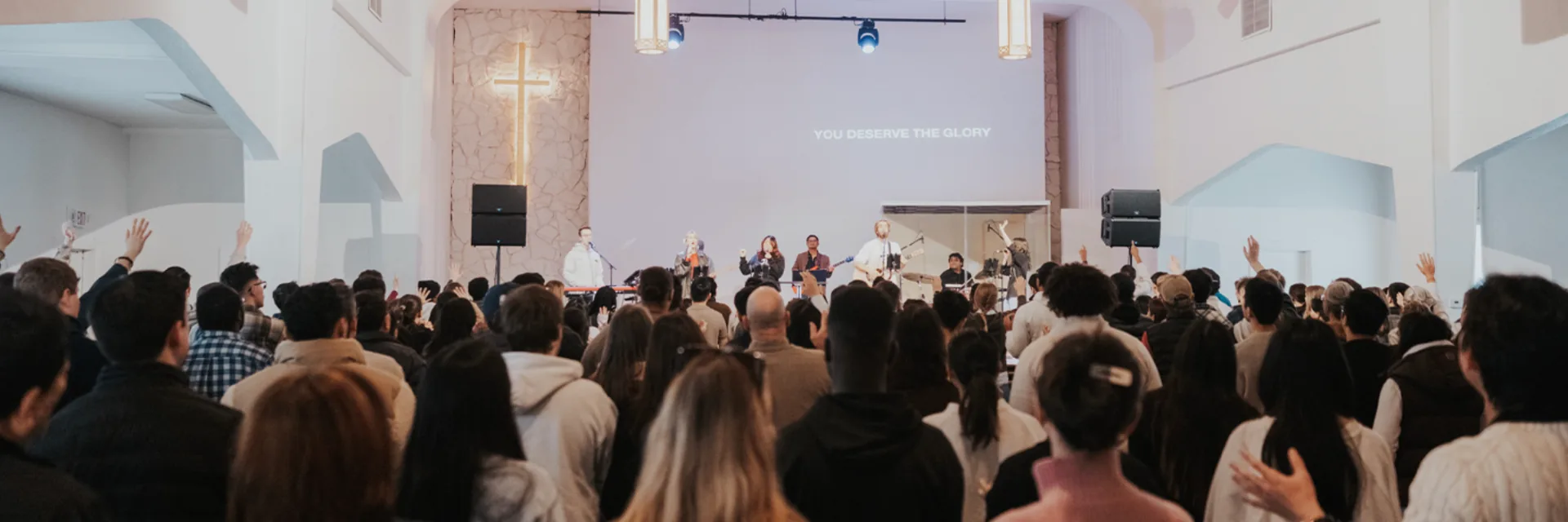 A congregation worshipping together in a church sanctuary, hands raised, with an illuminated cross on the wall